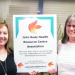 Chantelle (left) and Tracey (right) holding up a John Rudy Health Resource Centre Association banner