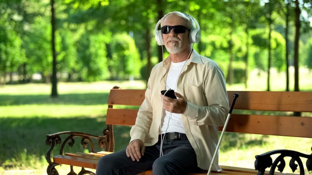 Man wearing dark glasses seated at a park bench. He is using headphones with his phone