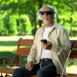 Man wearing dark glasses seated at a park bench. He is using headphones with his phone