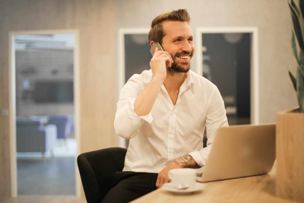 man speaking on phone, seated at table with his laptop