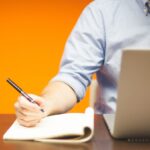 man at a laptop, writing in a notebook