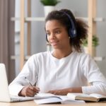 student using headphones and looking at laptop, taking notes in notebook