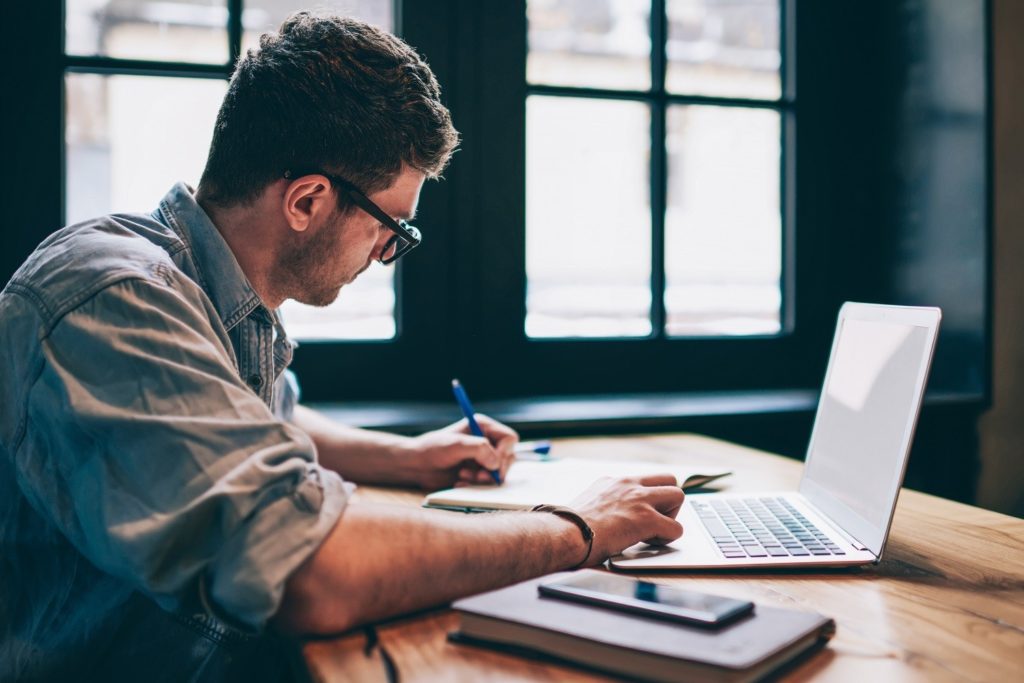 man using a laptop while writing in a notebook