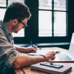 man using a laptop while writing in a notebook