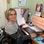 Lori working at her desk, seated in her wheelchair, looking at the camera and smiling