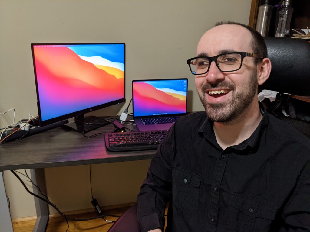 Nate with the equipment he received. He is smiling, seated in an ergonomic chair. Behind him is his desk, with a laptop, keyboard and external monitor