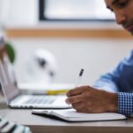 man working at a laptop, writing notes in a book