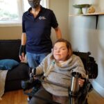 Marc stands next to Bonnie, who is using her wheelchair lap tray to hold a mug.