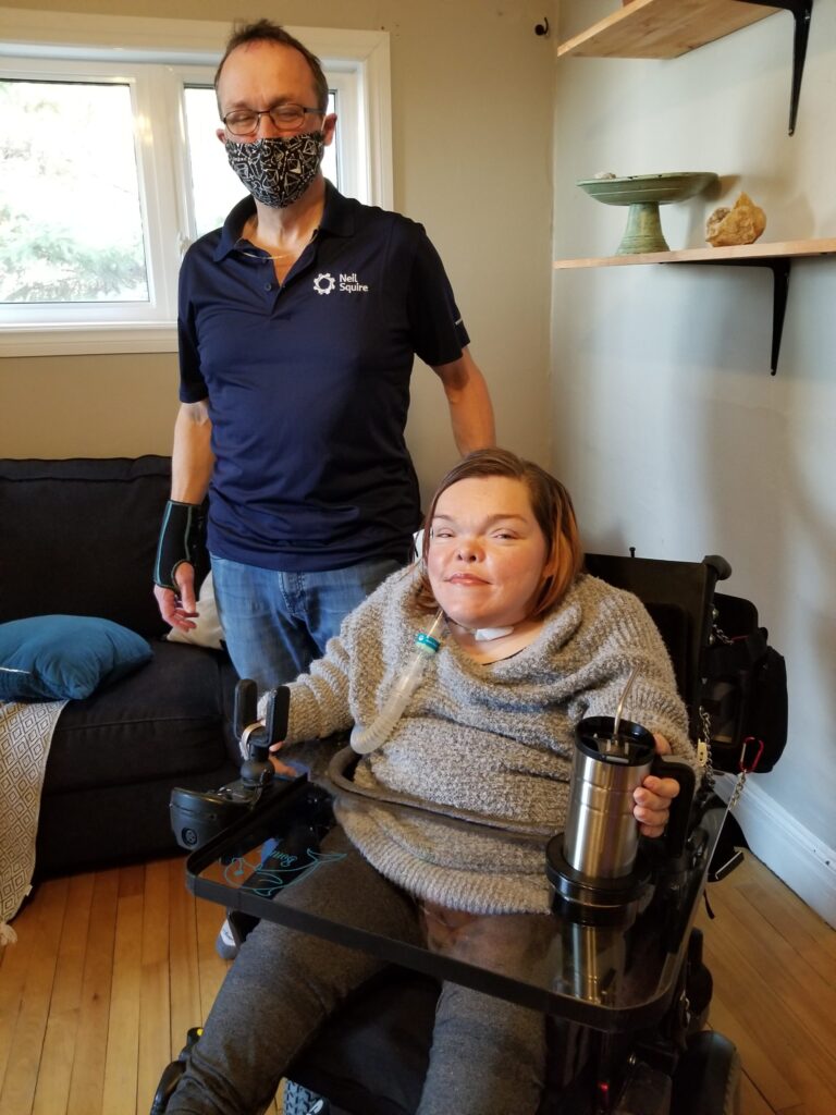 Marc stands next to Bonnie, who is using her wheelchair lap tray to hold a mug.