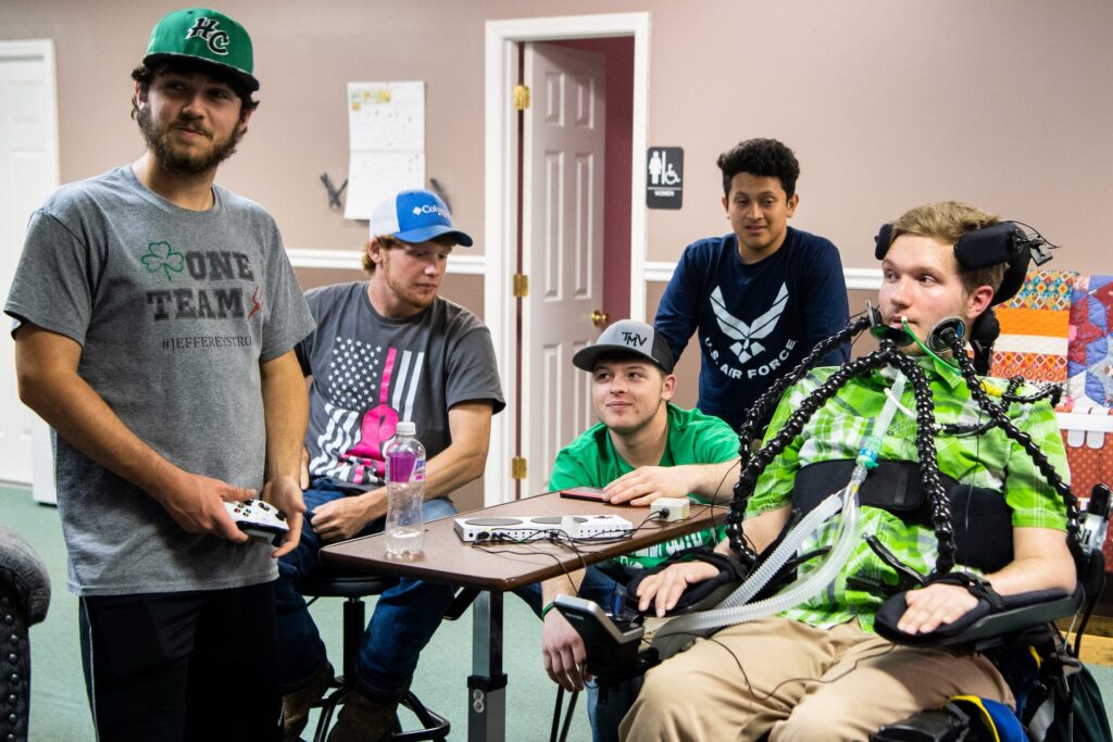 A man in a wheelchair plays video games with friends using assistive technology.