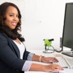 A woman working at a desk, using a keyboard and mouse. She is smiling at the camera