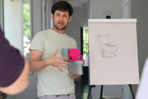 Noam Platt is talking while holding up a small toy-sized toilet, standing in front of a design for a toilet.