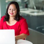 Woman wearing red, seated at a desk, typing on a laptop