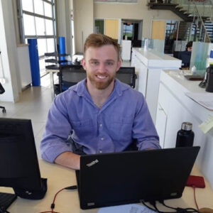 Working Together participant Blake sitting at his desk at work, smiling and working on his laptop