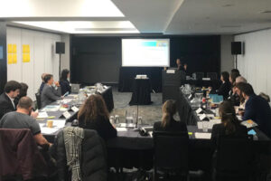 A networking session with people sitting at a long table, watching presenter and a PowerPoint presentation