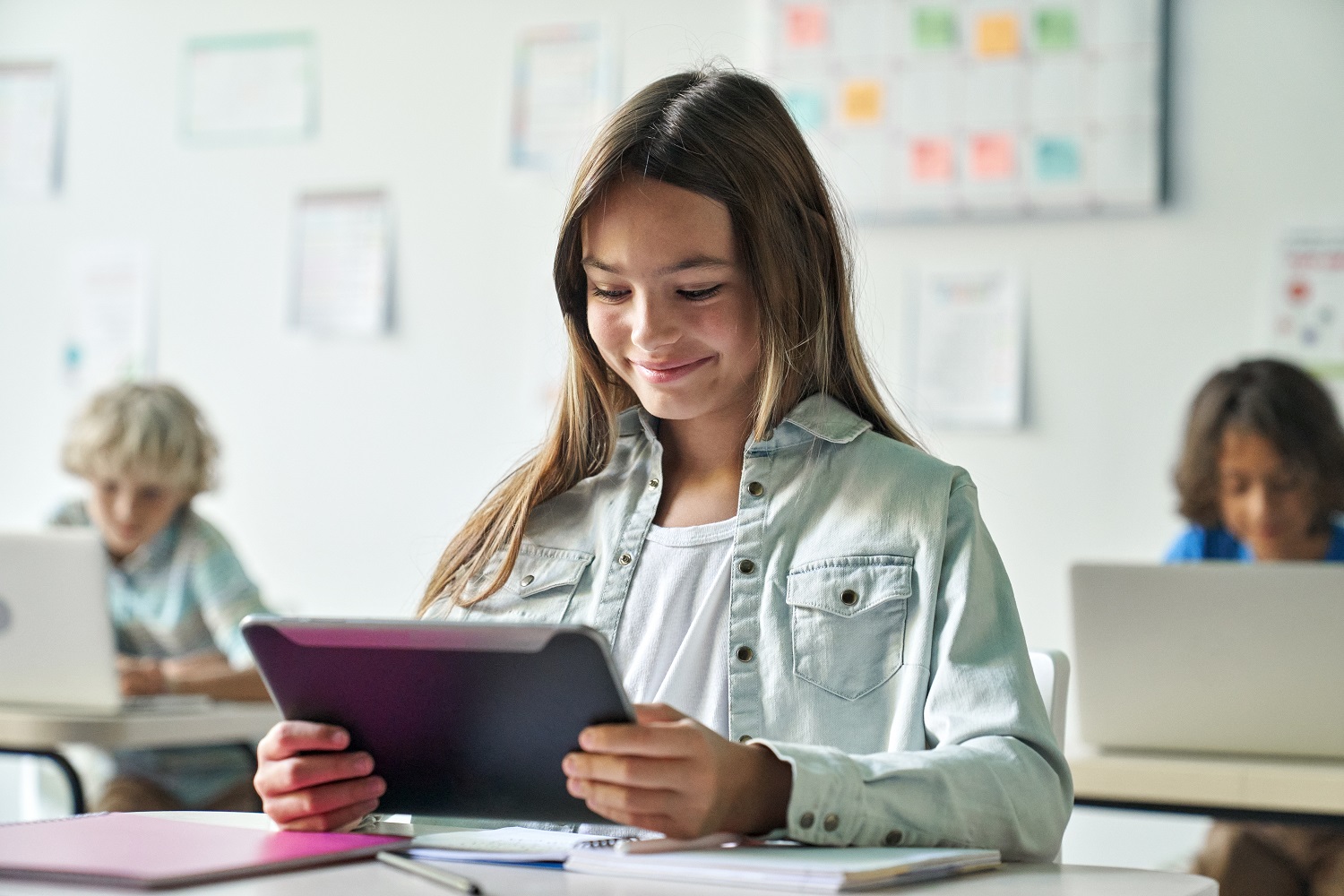 A young girl in a classroom smiles while using a tablet.