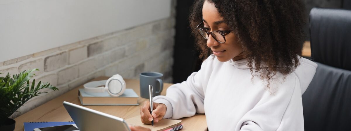 A woman writes on a notepad while watching something on a tablet.