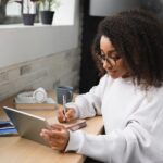 A woman writes on a notepad while watching something on a tablet.