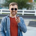 A blind man wearing sunglasses and holding a cane, speaks on the phone.