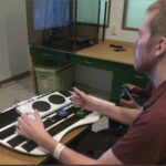 A man in a wheelchair using the Xbox Adaptive Controller and assistive switches to play video games.