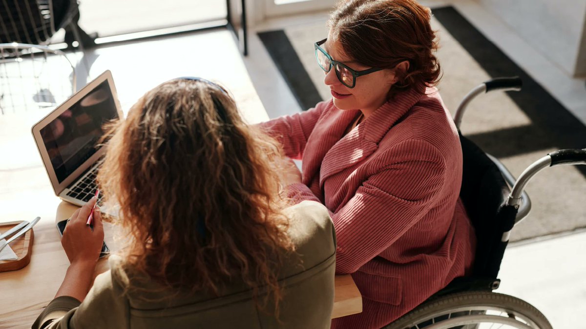 A woman in a wheelchair talks with another woman, while they look at a laptop