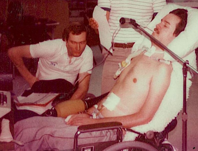 Gary Birch, in a wheelchair, looks over Neil, in a bed with a sip and puff device mounted.