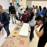 A busy room, as university students work using cardboard, Styrofoam, and other common materials to make devices.