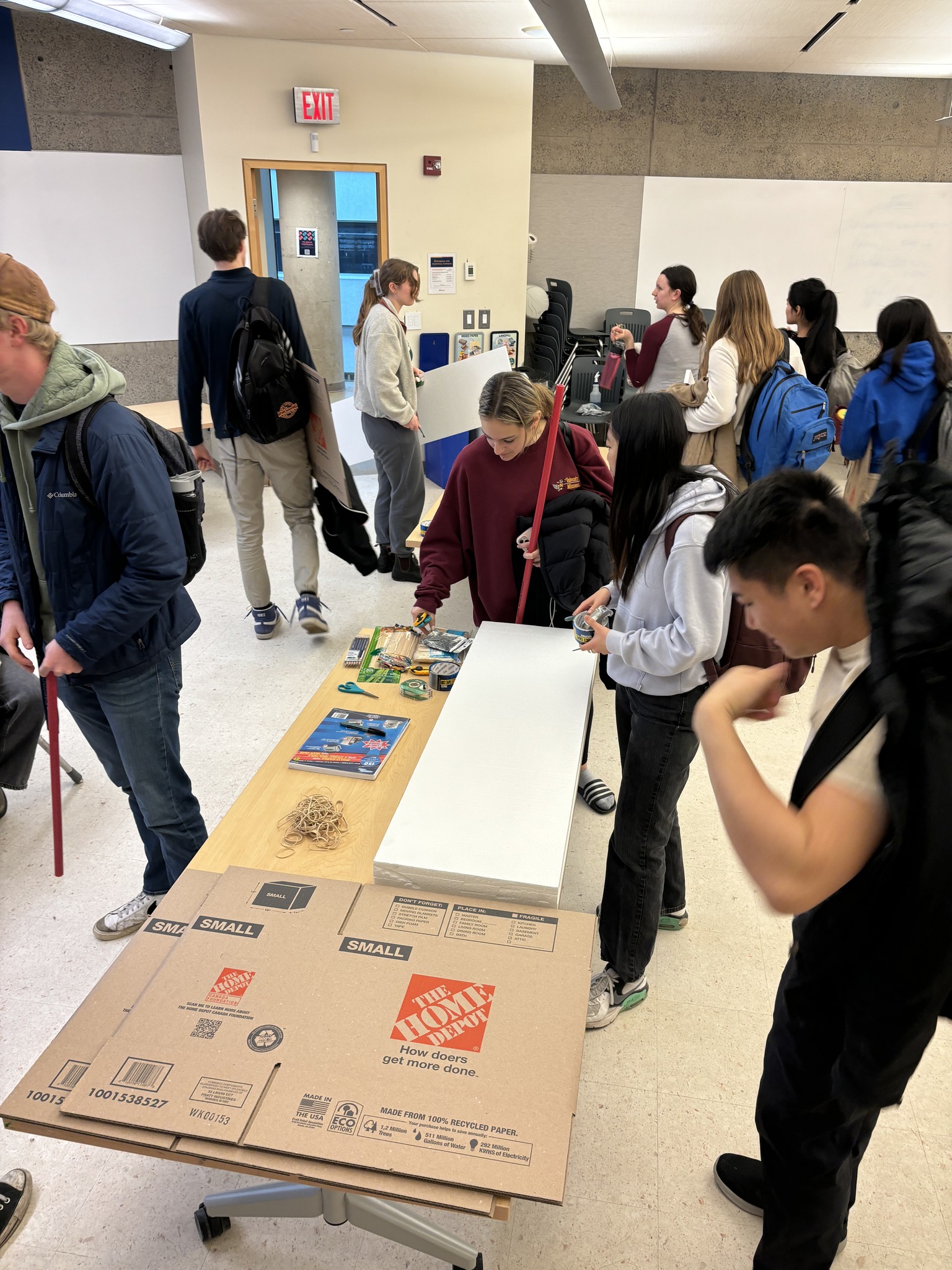 A busy room, as university students work using cardboard, Styrofoam, and other common materials to make devices.