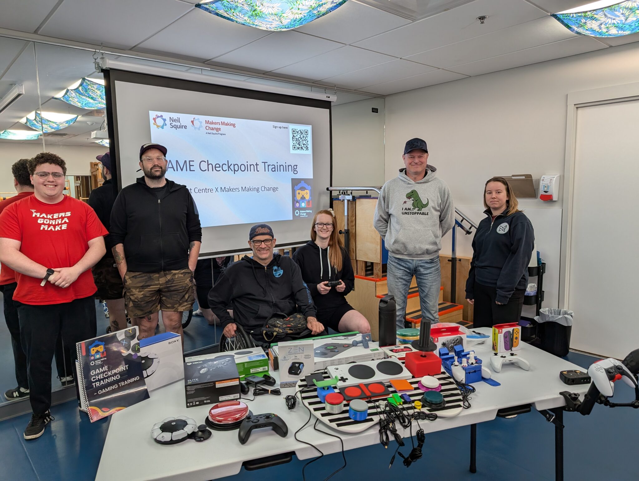 MMC Accessible Gaming Lead Tyler Fentie, Movement Centre Lead Conductor Chris Martin, Brandon chapter leader Russ Mitchell, and Movement Centre staff sit behind a table with Adaptive Gaming equipment during training.