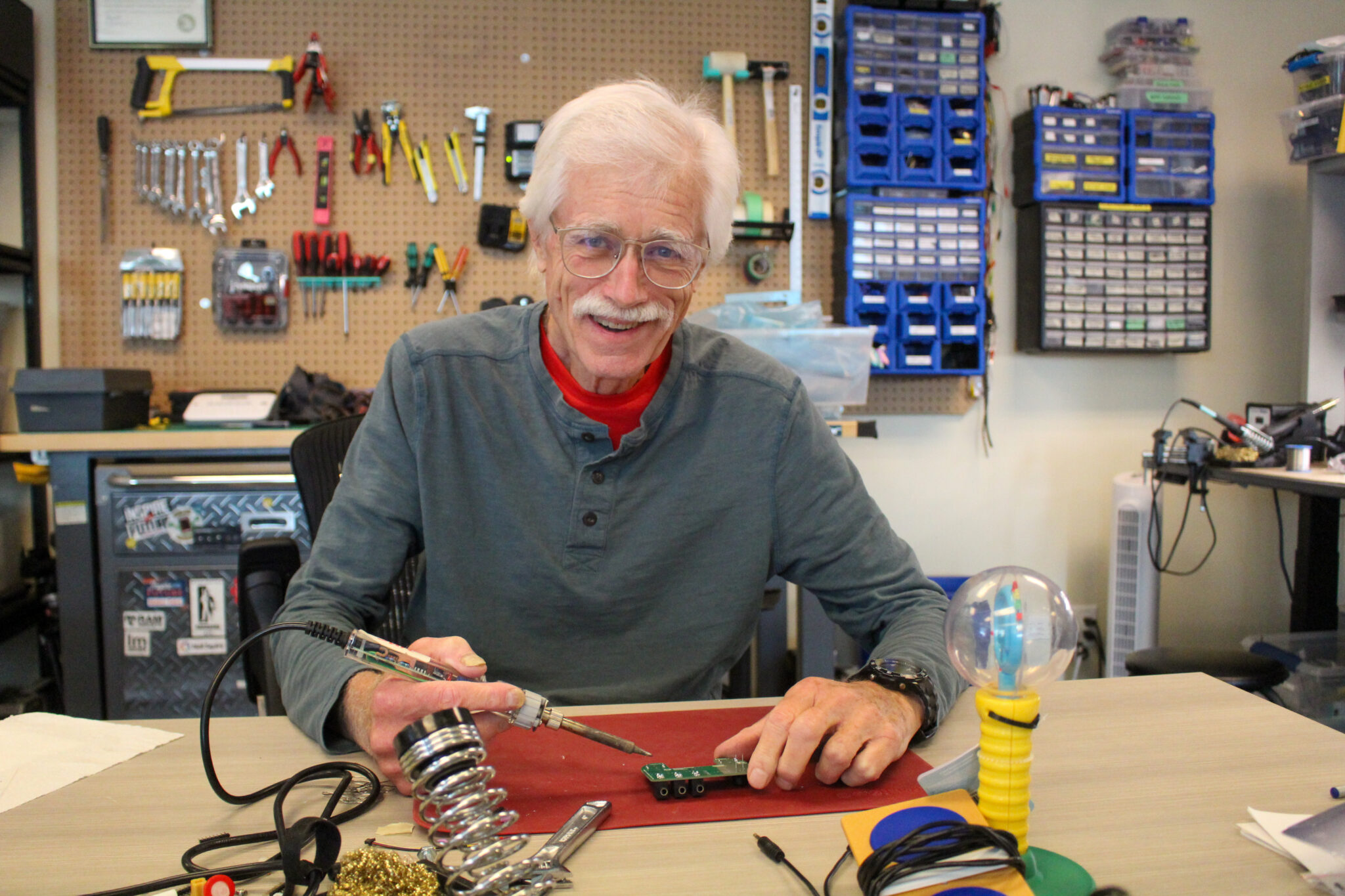 Bob smiles while holding a soldering iron to work on a device, with a backdrop of tools on the wall behind him.