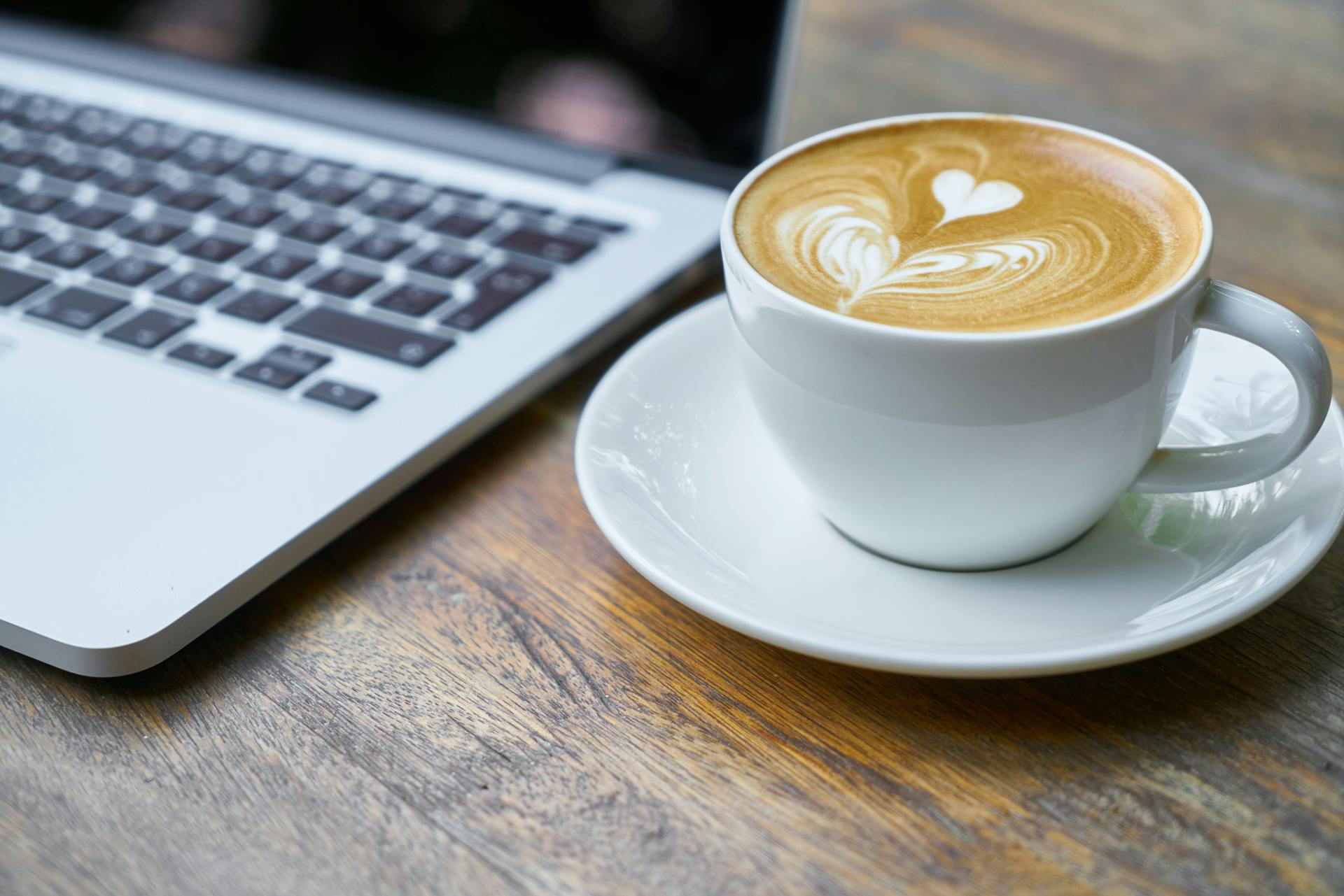 A cup of coffee with the foam made into the shape of a heart next to a laptop on a table.