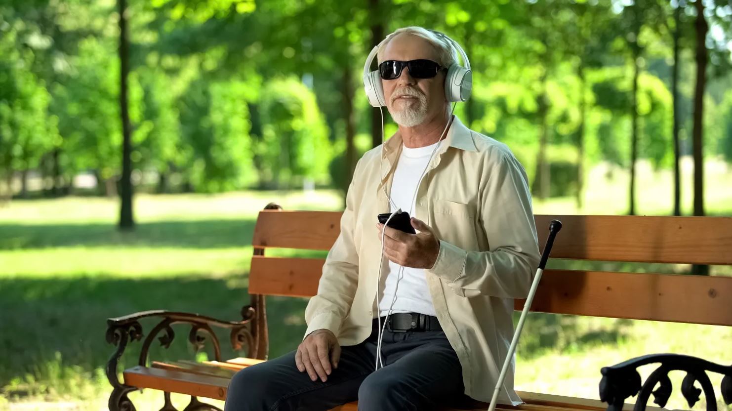 A blind man wearing sunglasses sits on bench wearing headphones and holding his phone.