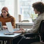 A woman in a wheelchair uses a laptop at a table, sitting close to a man also using a laptop.