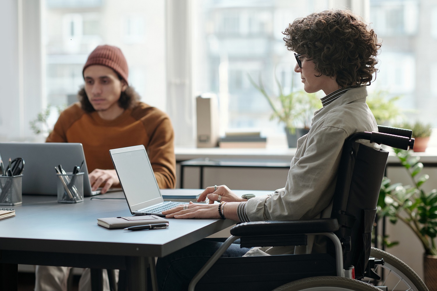 A woman in a wheelchair uses a laptop at a table, sitting close to a man also using a laptop.