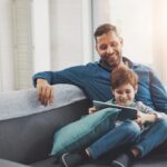 A father smiles as he watches his son use a tablet.