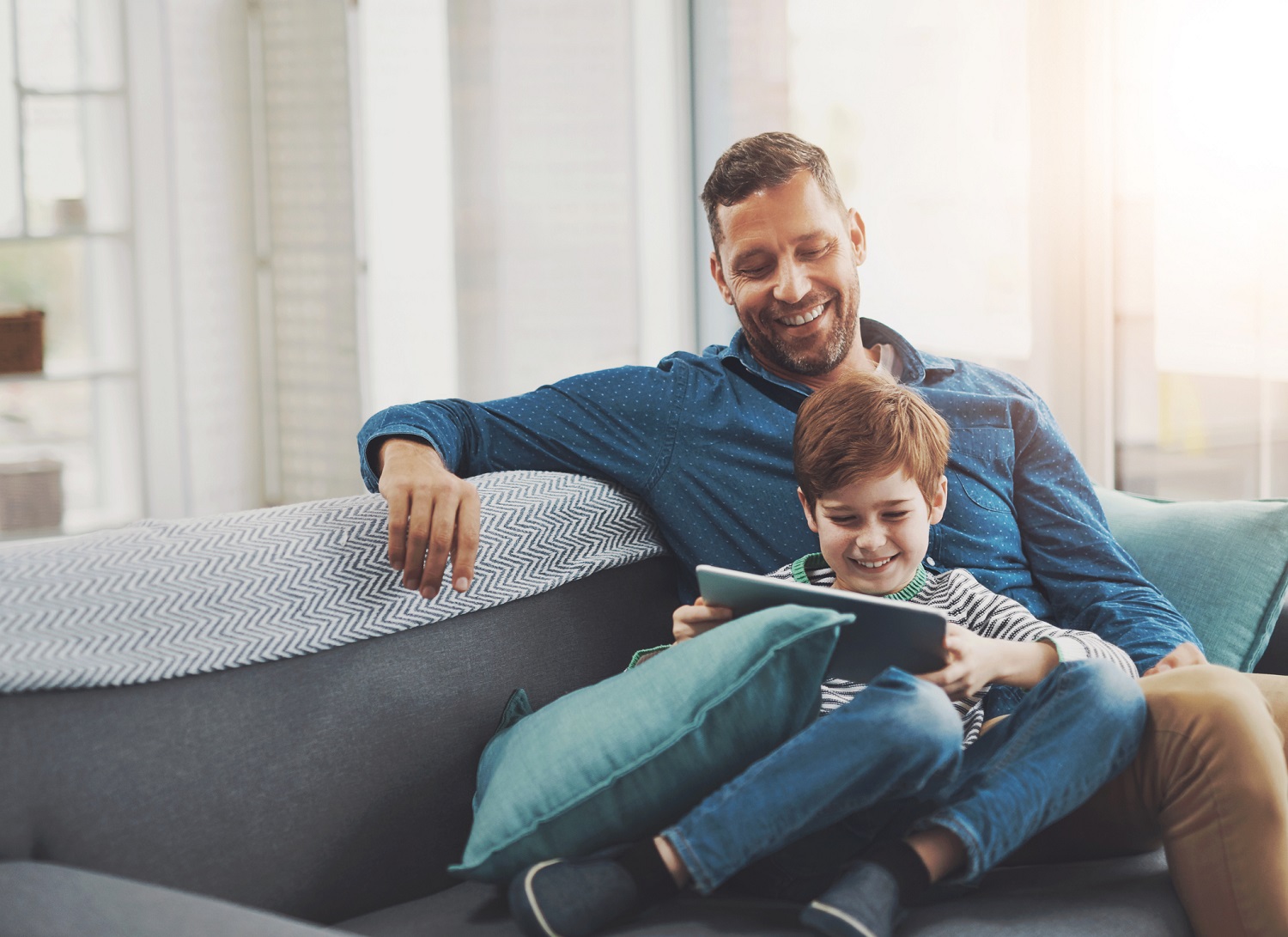 A father smiles as he watches his son use a tablet.