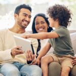 Parents sit with their child on a couch, using a tablet together as they all smile.