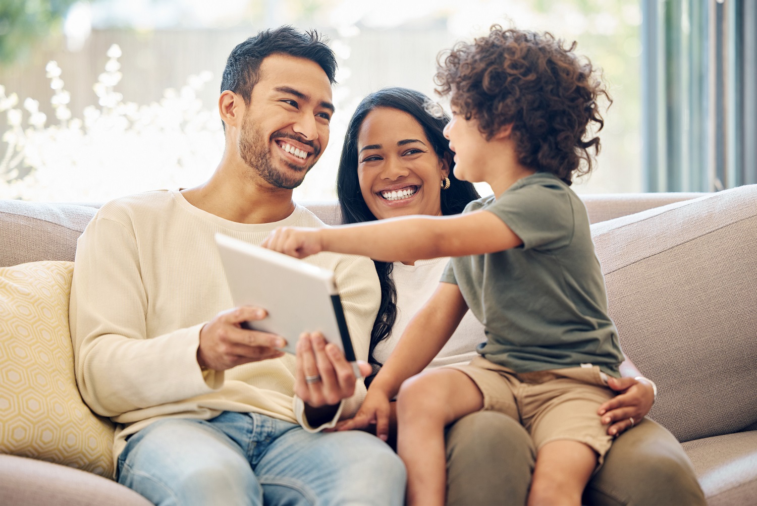 Parents sit with their child on a couch, using a tablet together as they all smile.